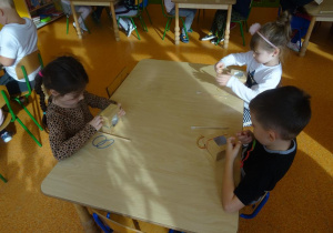 Troje dzieci siedzi przy stole, w rękach trzymają gumki recepturki, które zakładają na kartonowe małe pudełko.